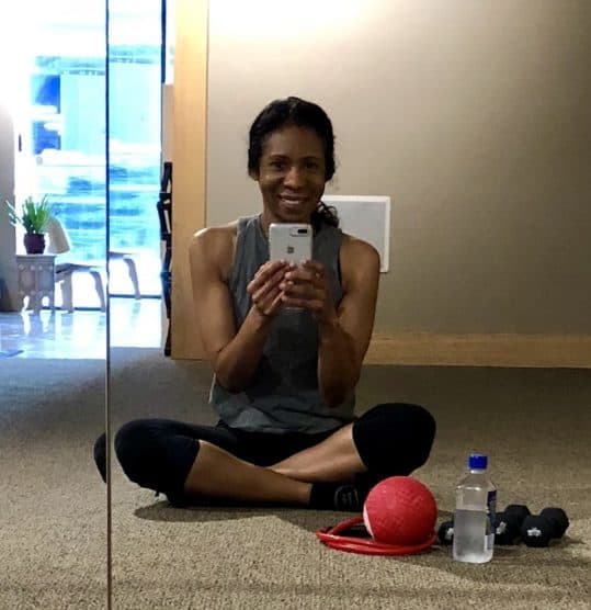 Sitting cross-legged on the barre studio floor in a pair of cropped black leggings and a gray muscle tee