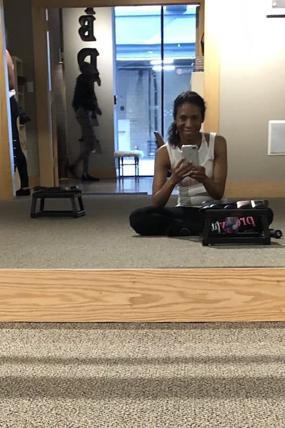 sitting crossed leg in the barre studio in a pair of black leggings and a white tank top
