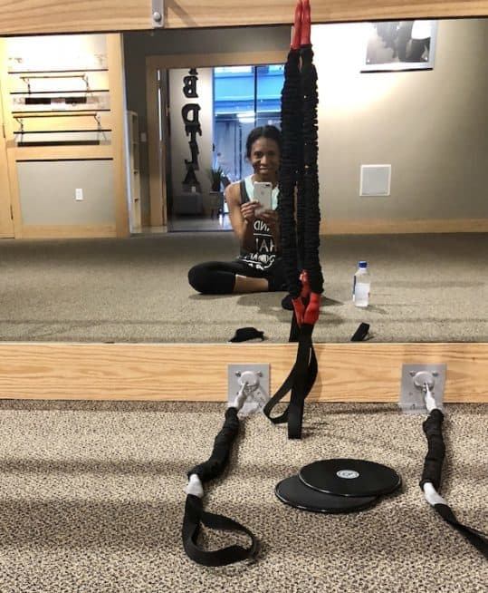 sitting cross legged in the barre studio in front of resistance bands and sliders.  I am wearing black cropped leggings and a gray tank.