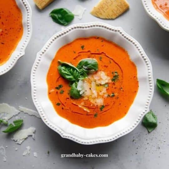 a white bowl of homemade tomato soup garnished with shaved cheese and basil leaves