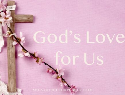 A wooden cross is laying on a pink table that is partially covered by a white lace table covering. A vine with pink flowers is laying across the top of the cross and it has a sign on the the top with the letters INRI