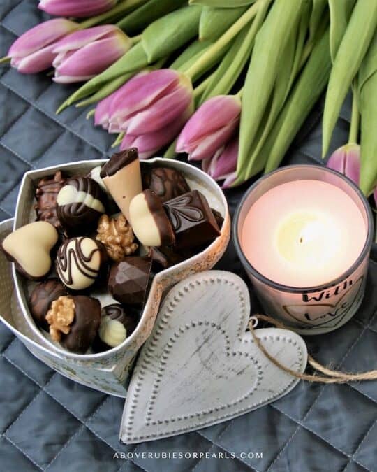 a heart shaped box of chocolates is displayed next to a let candle that says "with love" and a bouquet of pink flowers