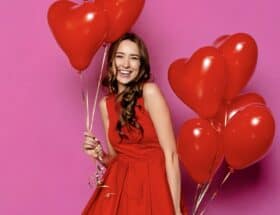 a smiling lady in a red sleeveless party dress is holding 8 heart-shaped red balloons