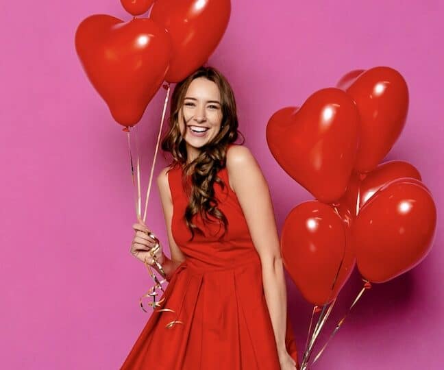 a smiling lady in a red sleeveless party dress is holding 8 heart-shaped red balloons
