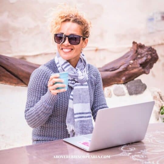 A smiling Proverbs 31 Business Woman working at her laptop outside. She is wearing sunglasses, a sweater, and a scarf.