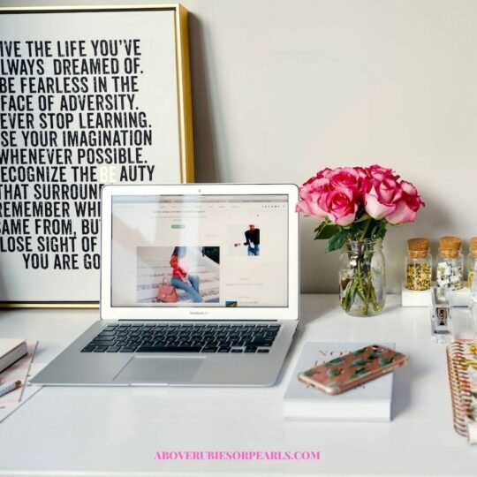A Proverbs 31 Business Woman home office. There is an open laptop on a white desk. Next to it is inspirational artwork encouraging her to live the life she always dreamed of. On the other side of it is a bouquet of pink roses. Also on the desk are books, a smart phone, a spiral notebook, and desk accessories.