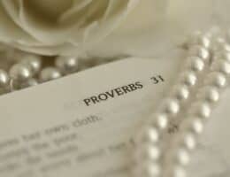 A Bible is open to a scripture describing the Proverbs 31 Woman. A string of pearls is on top of the Bible and it is open next to a rose.