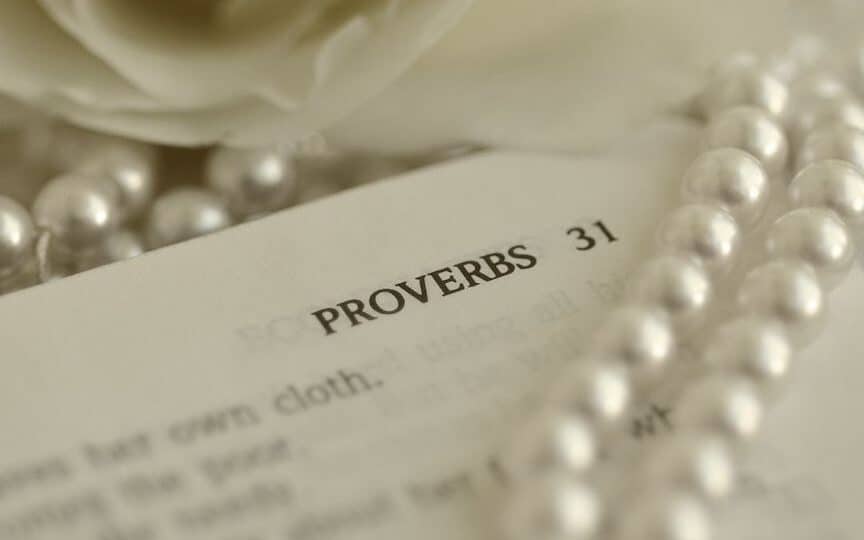 A Bible is open to a scripture describing the Proverbs 31 Woman. A string of pearls is on top of the Bible and it is open next to a rose.