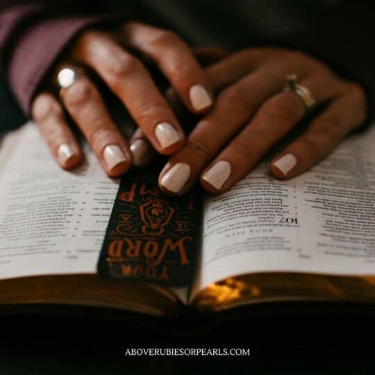A lady's lovely hands on gracefully placed on top of an open Bible. There is a bookmark holding her place. Her nails are neatly manicured and painted a pearly white. She is wearing her engagement ring and wedding band on one hand and another ring on her other hand.
