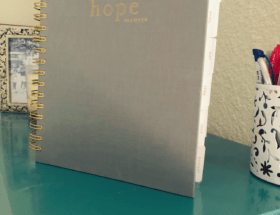 A spiral-bound planner is standing on a teal-colored table. It is gray with gold lettering that says, "Hope Planner." A bookmark labeled, "Today," is sticking out of the top. It is in front of a candle and a silver picture frame. Next to it is a white metal pencil put full of pens. The pencil cup has various shapes cut out of it.