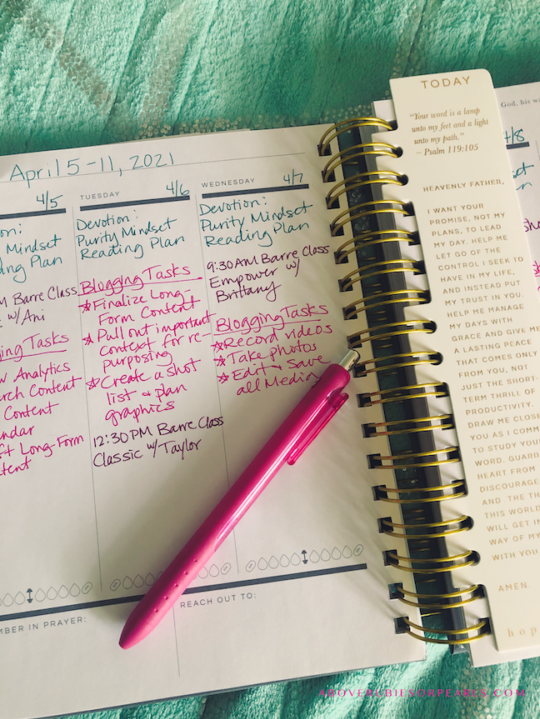 A spiral-bound planner is open on a bed covered in a teal blanket with silver accents. On the left is a page that shows a week at a glance. On the right is a bookmark labeled, "Today." The bookmark also contains a scripture and a prayer. The week at a glance page is filled in with to-do lists in different colored ink.