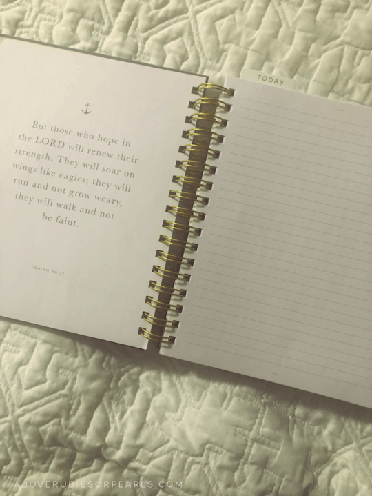 A spiral-bound planner is open on a bed with a white quilted bedspread. There is a scripture on the left side and a lined page on the right to take notes.