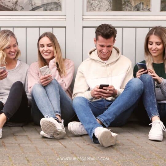A clique of three girls and one boy are sitting on the ground, laughing at something on their phones.