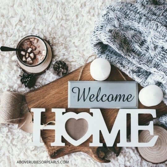 A cozy throw, a cup of hot chocolate, and candles are on a cozy rug with a 'welcome home' sign.