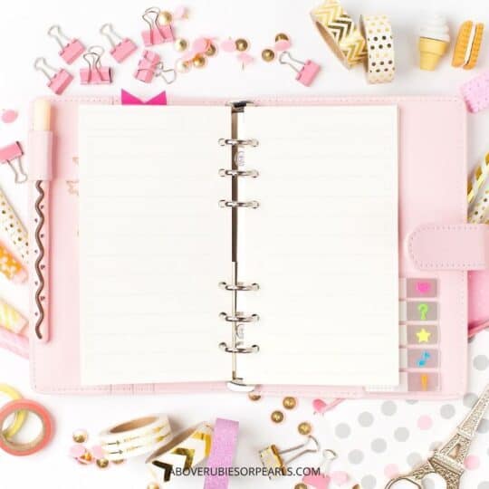 A pretty pink planner is opened on a desk with a display of pink binder clips, polka dotted pins, and rolls of color tape, stickers, and magnets all around it.