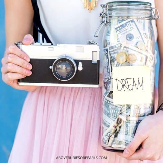 A lady is building her Proverbs 31 Woman home by holding a glass jar full of money she has saved in one hand with a label on it that says 'dream'. In the other hand, she is holding a camera.