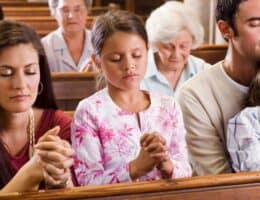 A Proverbs 31 family is sitting together at church. Their eyes are closed and their hands are clasped in prayer. The daughter is sitting between both parents and the son is sitting on the father’s lap.