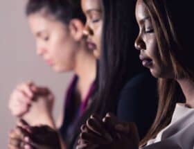 3 women are standing together in prayer with eyes closed and hands clasped