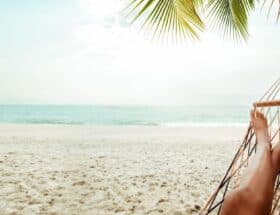 A lady is enjoying God's rest by relaxing in a hammock on the beach under a palm tree.