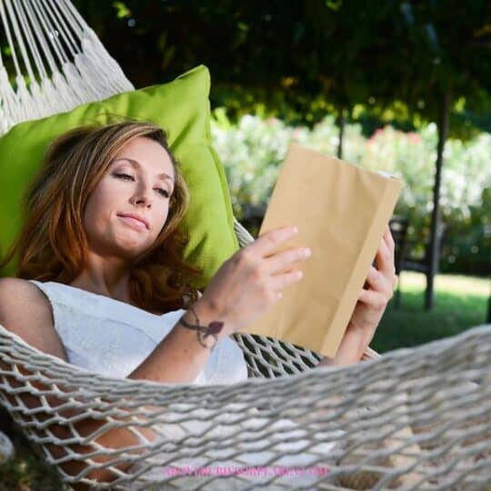 A lady is reading in a hammock with a chartreuse green pillow behind her head.