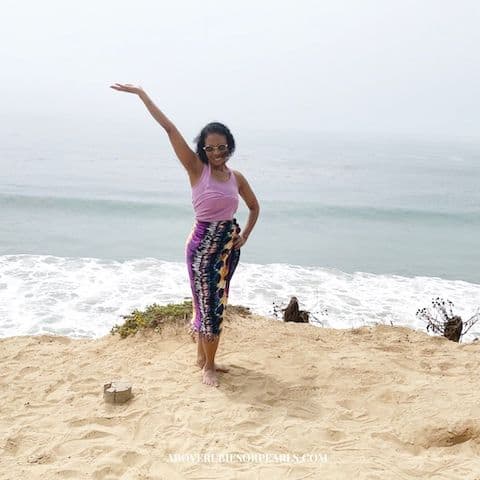 I am standing on the bluff above El Matador State Beach with one hand in the air and one hand on my hip. The beach is in the background and the wind is whipping my hair. I am dressed in a tank top and a sarong.
