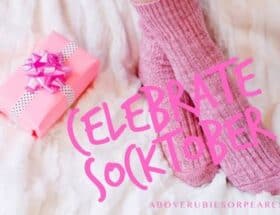 A pair of feet clothed in a pair of pink socks are resting on a soft, cozy rug and next to a gift box wrapped in pink paper, tied with a white and black polka dot ribbon and with a pink shiny bow on top.