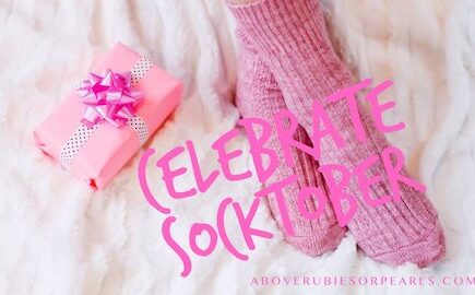 A pair of feet clothed in a pair of pink socks are resting on a soft, cozy rug and next to a gift box wrapped in pink paper, tied with a white and black polka dot ribbon and with a pink shiny bow on top.