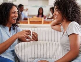 An older and a younger woman chat on a sofa. The older woman is holding a cup of coffee. The younger woman is smiling. The older woman is laughing.