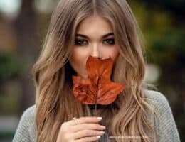 A lady in modest fashion is covering her mouth with a fall leaf.