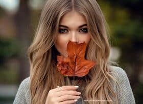 A lady in modest fashion is covering her mouth with a fall leaf.