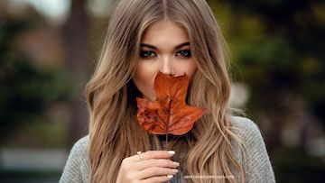 A lady in modest fashion is covering her mouth with a fall leaf.