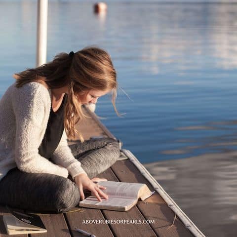 A lady is sitting cross-legged on the pier reading the Bible.