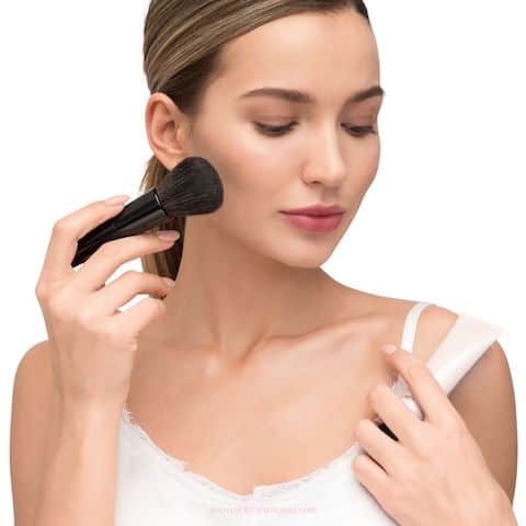 Lady in white dress applying blush to cheek with makeup brush