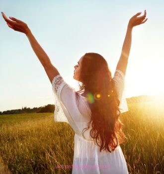 Lady in a white dress standing in a field of wheat with arms extended in the air while the sun is rising