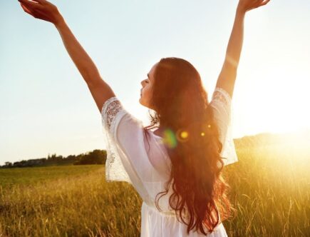 Lady in a white dress standing in a field of wheat with arms extended in the air while the sun is rising
