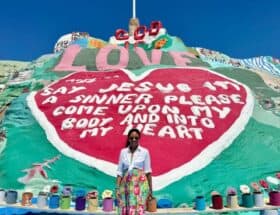 Woman standing in front of Salvation Mountain in the California desert with the painted “God Is Love” message and cross visible above.