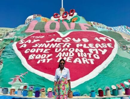 Woman standing in front of Salvation Mountain in the California desert with the painted “God Is Love” message and cross visible above.