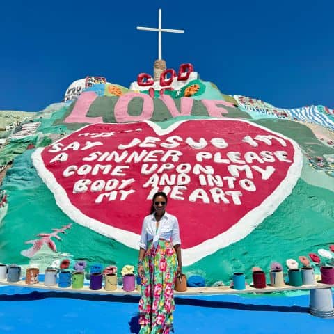 Woman standing in front of Salvation Mountain in the California desert with the painted “God Is Love” message and cross visible above.