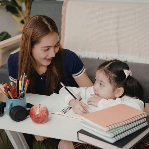 Woman helping a young girl with schoolwork at a table filled with pencils and a notebook, illustrating generosity through giving time