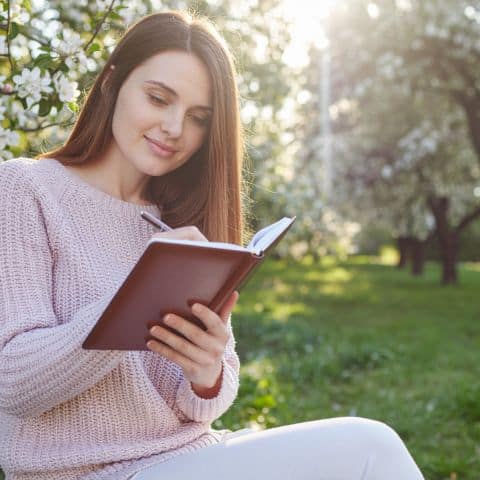 Woman journaling outdoors in spring sunlight beneath blossoming trees during a quiet moment of reflection