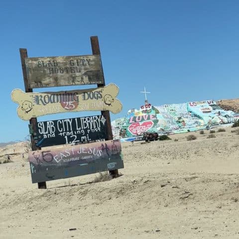Handmade wooden road signs in Slab City pointing to local landmarks like East Jesus Sculpture Garden and Slab City Library, with Salvation Mountain visible in the background.