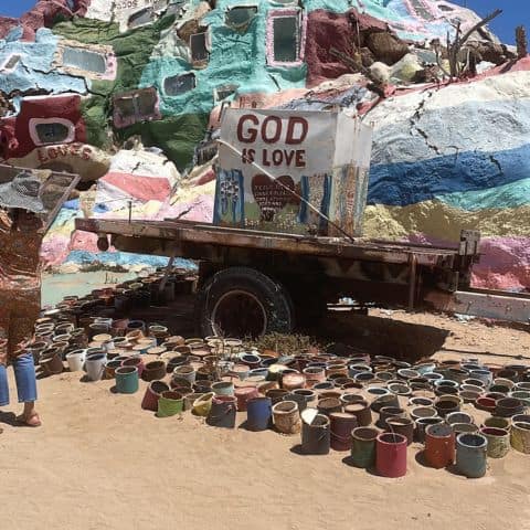Close-up of Salvation Mountain’s windowed structure with a “God Is Love” sculpture made from truck parts and colorful paint cans in front.