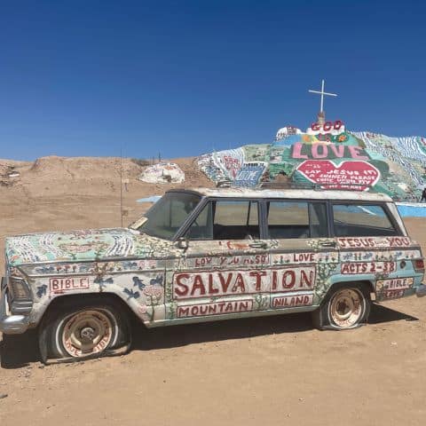 Painted car at Salvation Mountain covered in colorful messages including “Jesus Loves You” and “Acts 2:38,” with the mountain in the background.