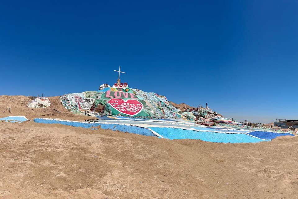Wide view of Salvation Mountain in the California desert, showing colorful artwork, painted Scriptures, and the cross on top.