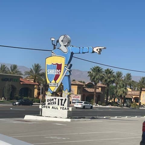 Giant knight statue in the parking lot of Shields Date Garden pointing toward the entrance with a shield-shaped sign reading “Shields” and “Don’t Miss It!”