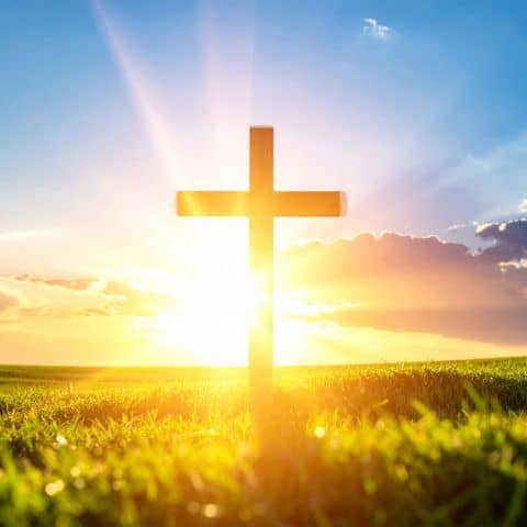 Wooden cross in a grassy meadow silhouetted by the sunrise, representing the hope and confidence found through the Cross.