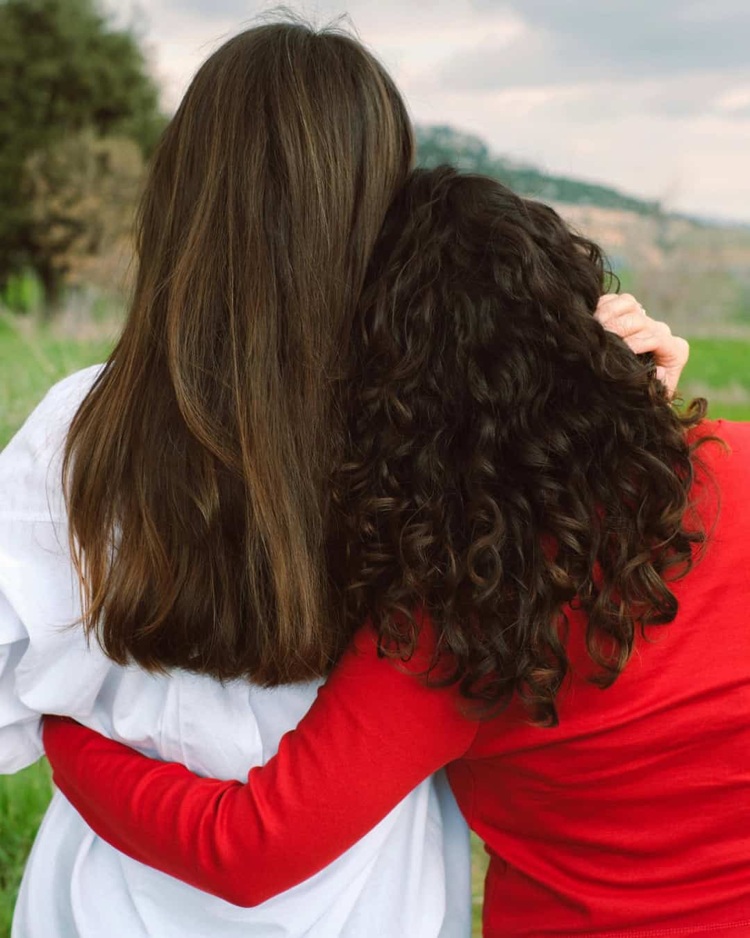 Two women embracing in a natural setting, one resting her head against the other, with trees and rolling hills in the background.