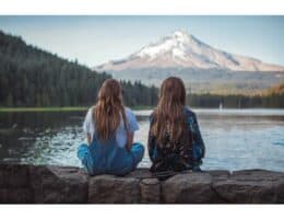 Two women sitting beside a lake, looking toward snow-capped mountains and an evergreen-covered mountainside.