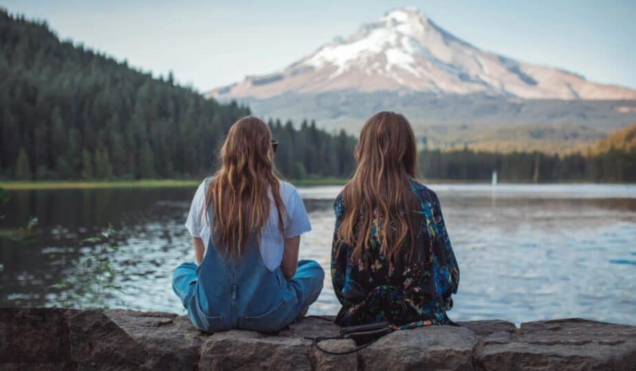 Two women sitting beside a lake, looking toward snow-capped mountains and an evergreen-covered mountainside.