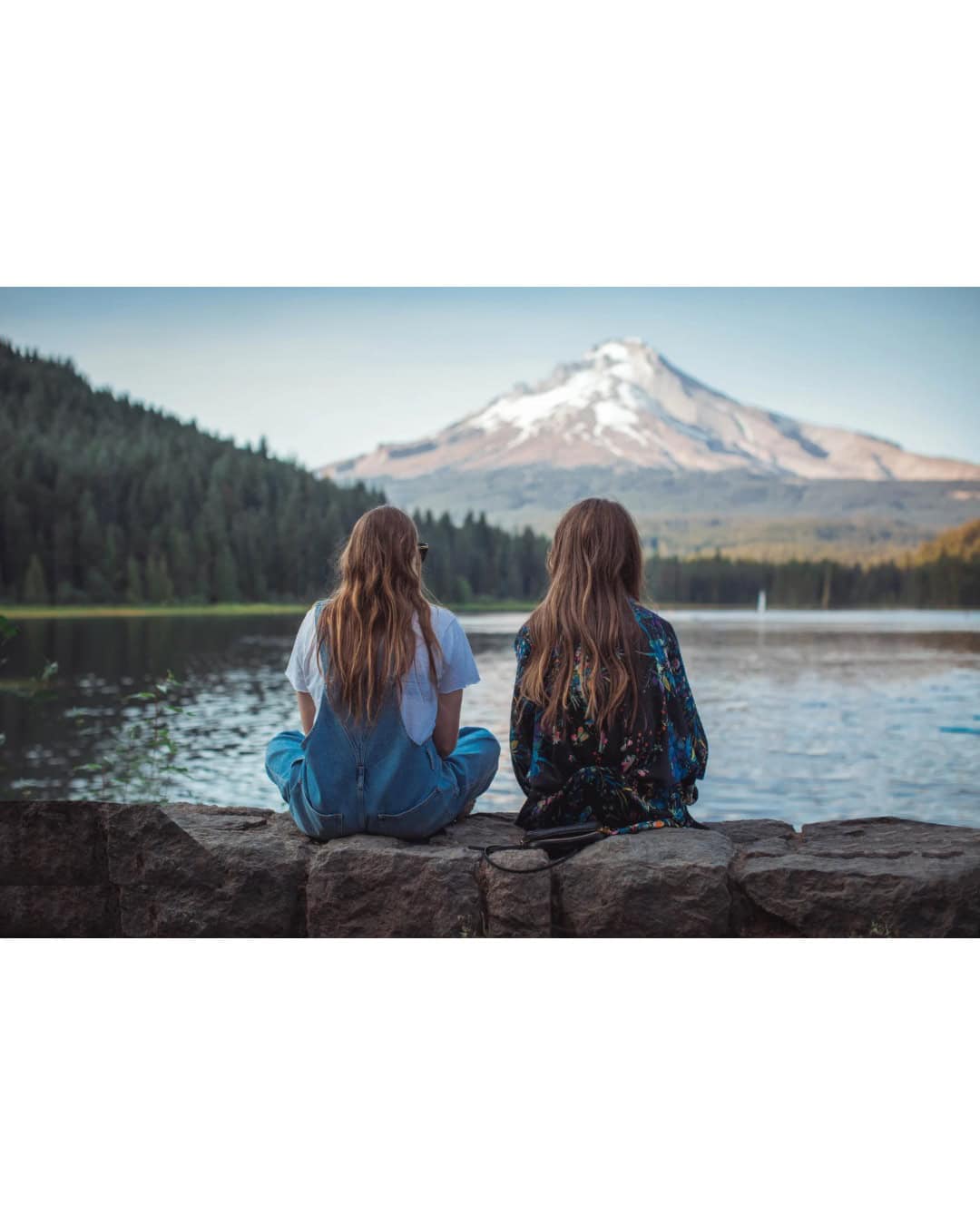 Two women sitting beside a lake, looking toward snow-capped mountains and an evergreen-covered mountainside.